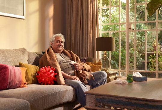 Homme âgé assis sur son divan; il a placé une boîte de mouchoirs sur sa table à café et une grande couverture sur ses épaules pour se réchauffer. Il semble présenter des symptômes de la COVID—19. À sa droite, un ballon qui représente le virus responsable de la COVID—19, hérissé de protéines Spike. L’homme regarde directement le lecteur, réfléchissant sciemment à son rendez-vous de vaccination contre la COVID—19.