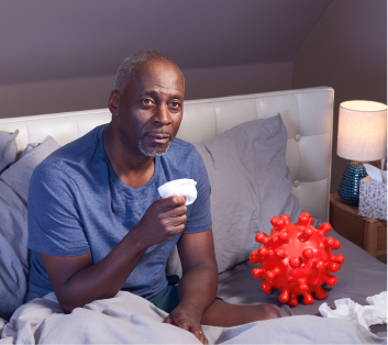 Adult patient sitting up in bed, holding a tissue in one hand. They appear to have symptoms ofCOVID-19. On theri left is a large replica of a COVID-19 Spike Protein ball. They are thinking about their COVID-19 high-risk factor