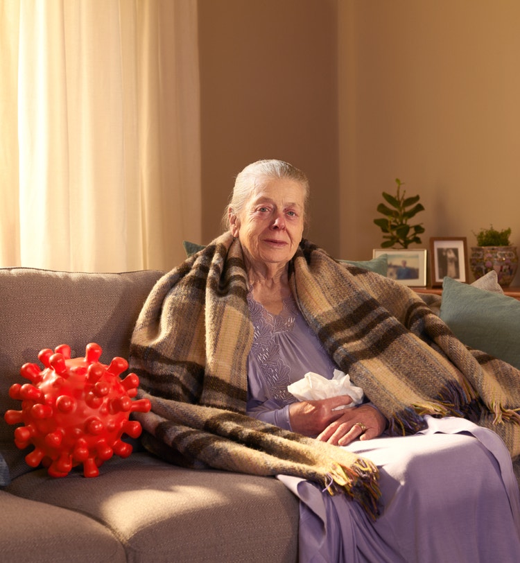 Older female patient sitting up on her home couch with tissues in her lap and a large blanket covering her shoulders to help keep warm. She appears to have symptoms of COVID—19. On her right is a large replica of a COVID—19 Spike Protein ball. She is looking directly at us, thoughtfully considering her COVID—19 high-risk factor