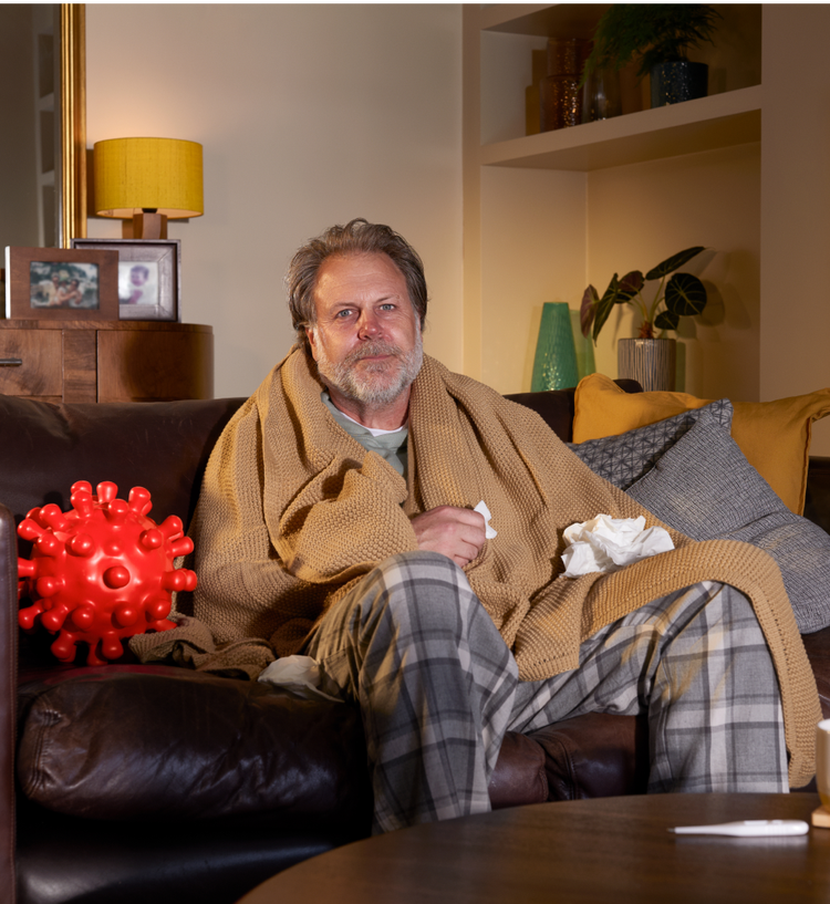 Older male patient sitting on his home couch with tissues in both hands. He is wearing a large, comfy bathrobe and pajamas. He appears to have symptoms of COVID—19. On his left is a large replica of a COVID—19 Spike Protein ball. He is looking directly at us, considering his COVID—19 high-risk factor.