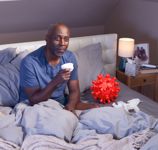 Adult patient sitting up in bed, holding a tissue in one hand. They appear to have symptoms of COVID-19. On theri left is a large replica of a COVID-19 Spike Protein ball. They are thinking about their COVID-19 high-risk factor.