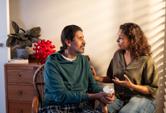 Two adults, one male patient and one female caregiver friend are having a discussion in a bedroom. The male patient is holding a tissue and seems to have what may be COVID—19 symptoms. They are talking about COVID—19 high-risk factors.