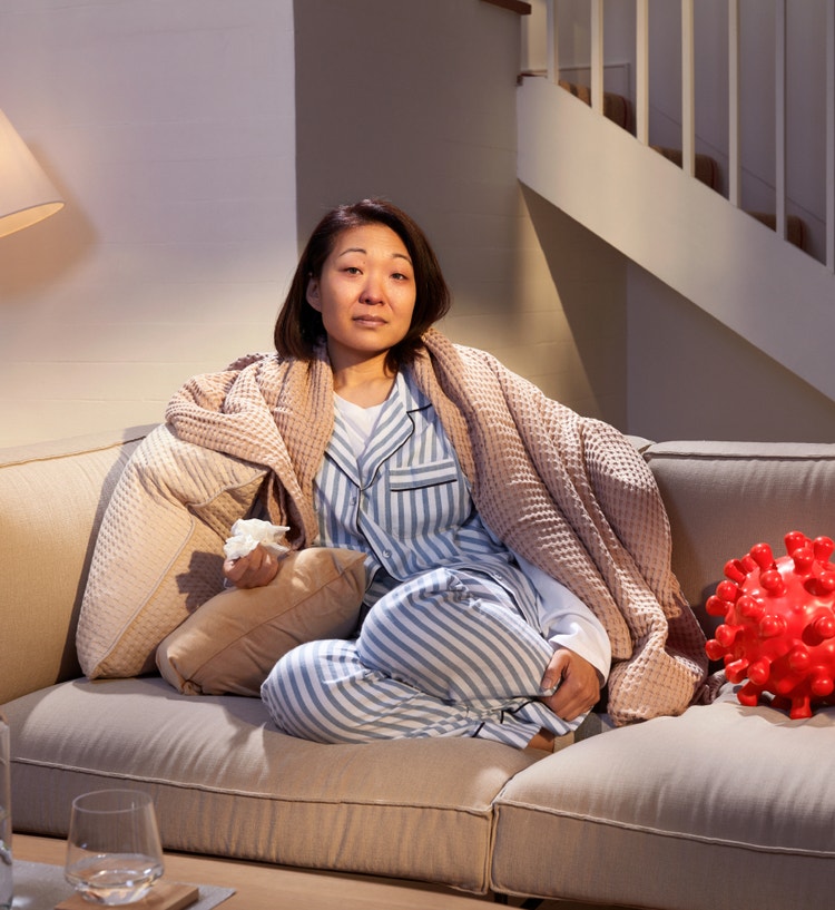 Adult female patient curled up on her home couch with a tissue in one hand and a large blanket covering her shoulders to help keep warm. She appears to have symptoms of COVID—19. On her left is a large replica of a COVID—19 Spike Protein ball. She is looking directly at us with a look of confidence related to her COVID—19 high-risk factor plan.
